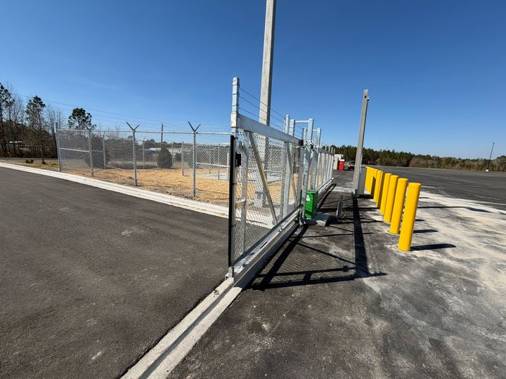 Galvanized chain-link commercial sliding gate with operator equipment, protected by yellow bollards.