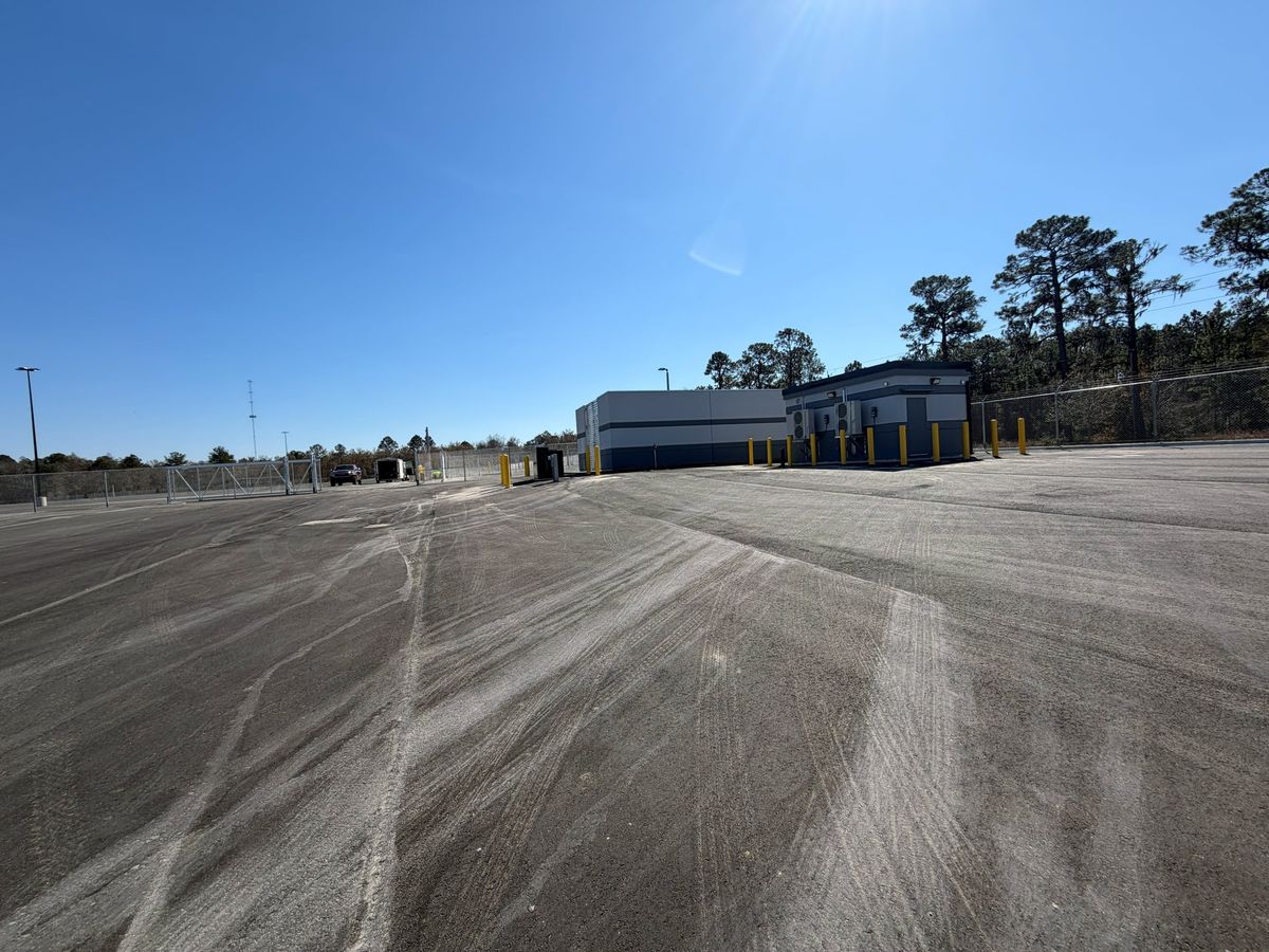 Large paved industrial site with perimeter chain-link fencing and a gate in the distance.