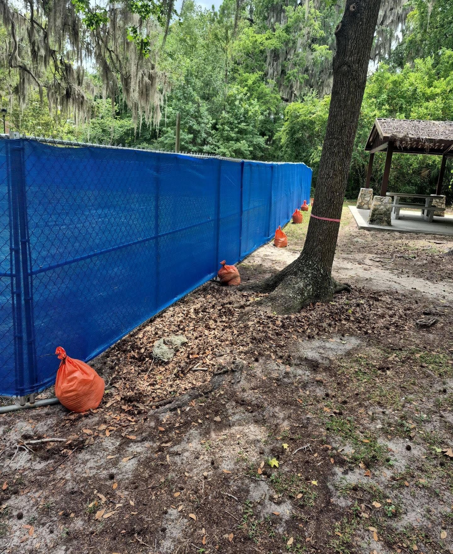 Blue privacy windscreen on a temporary chain-link fence, held down with ballast weights at a park perimeter.